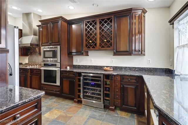 kitchen with wall chimney exhaust hood, stainless steel appliances, beverage cooler, and dark stone counters