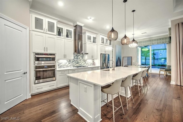 kitchen featuring appliances with stainless steel finishes, decorative light fixtures, white cabinets, wall chimney range hood, and a center island with sink