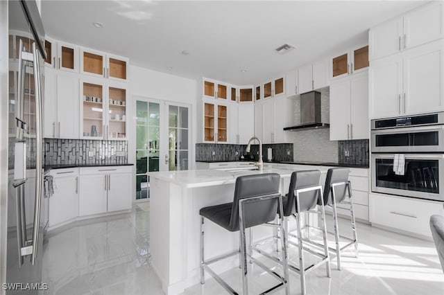 kitchen featuring wall chimney range hood, decorative backsplash, a kitchen island with sink, and stainless steel double oven