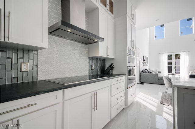 kitchen featuring black electric stovetop, a towering ceiling, wall chimney range hood, backsplash, and white cabinets