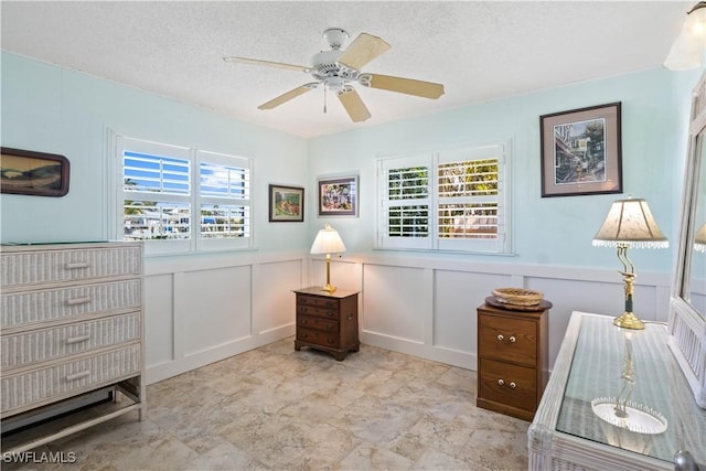 sitting room with a textured ceiling, ceiling fan, wainscoting, and a decorative wall