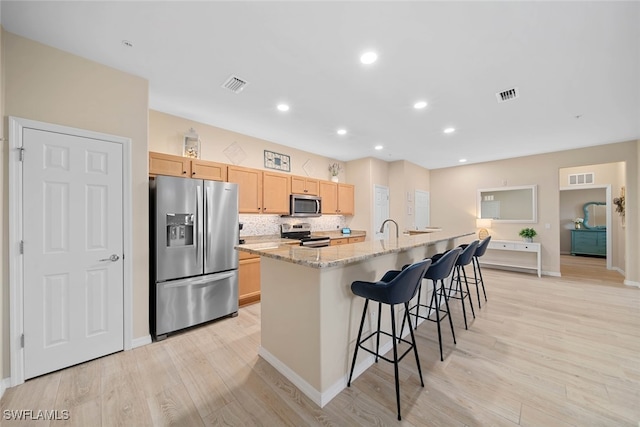 kitchen featuring light brown cabinets, an island with sink, light hardwood / wood-style flooring, stainless steel appliances, and light stone countertops