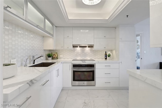 kitchen featuring stainless steel oven, sink, backsplash, a tray ceiling, and white cabinets