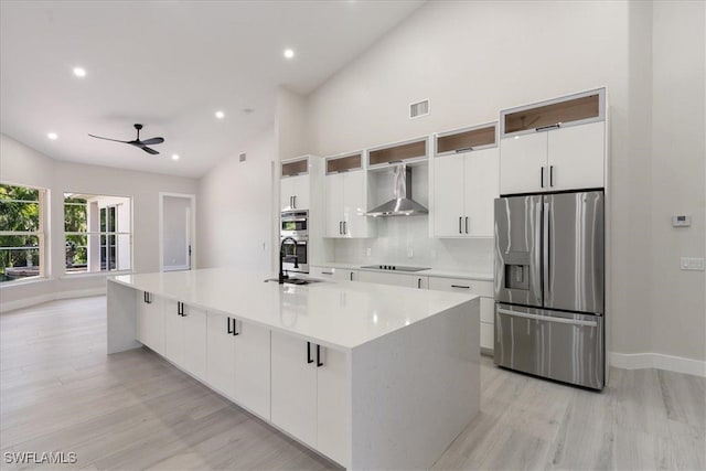 kitchen featuring wall chimney range hood, white cabinets, appliances with stainless steel finishes, a kitchen island with sink, and light wood-type flooring