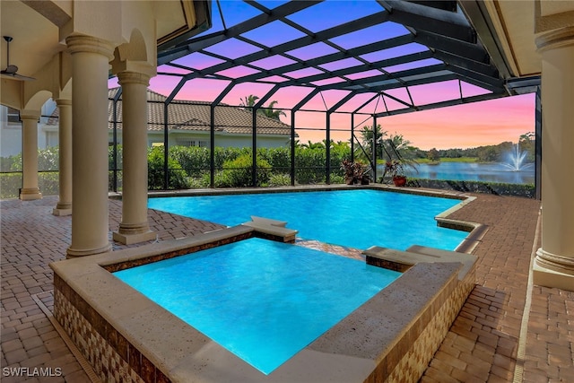 pool at dusk featuring an in ground hot tub, a water view, ceiling fan, a patio, and a lanai