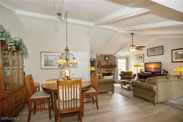 dining room featuring a brick fireplace, light hardwood / wood-style flooring, vaulted ceiling with beams, and ceiling fan with notable chandelier