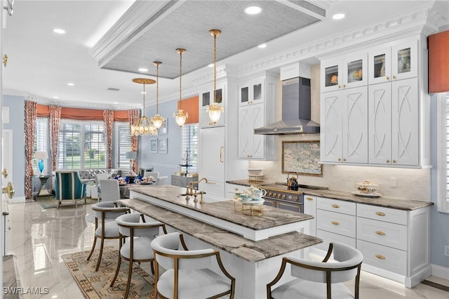 kitchen featuring white cabinetry, wall chimney range hood, dark stone countertops, an island with sink, and pendant lighting