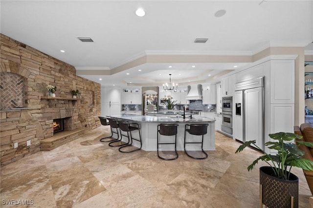 kitchen featuring paneled refrigerator, a fireplace, custom range hood, a large island with sink, and white cabinets