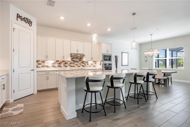 kitchen with under cabinet range hood, a kitchen island with sink, white cabinetry, and light countertops