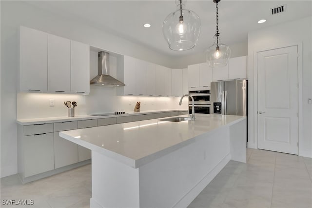 kitchen with wall chimney range hood, appliances with stainless steel finishes, an island with sink, and decorative light fixtures