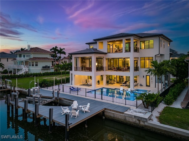 back house at dusk featuring a water view, a balcony, a patio area, and a pool with hot tub