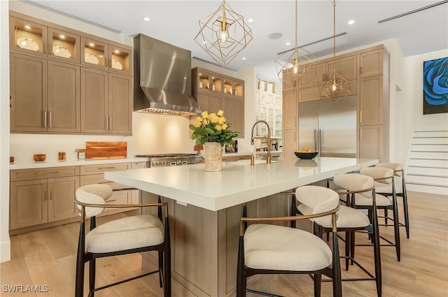 kitchen with pendant lighting, a large island with sink, built in refrigerator, wall chimney range hood, and light wood-type flooring