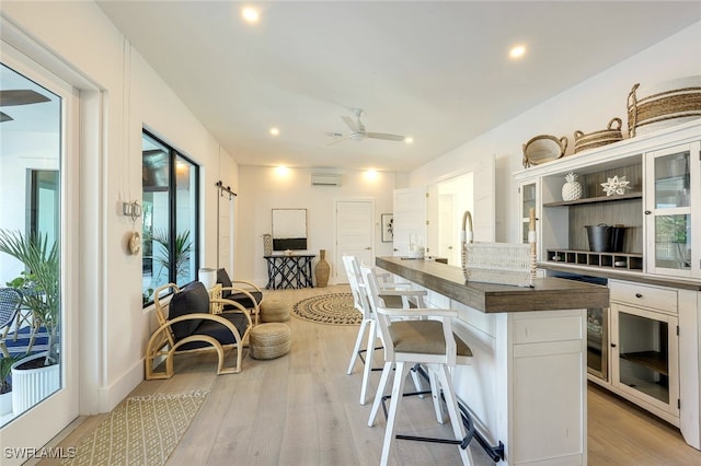 kitchen with a wall mounted AC, white cabinets, ceiling fan, a center island with sink, and light wood-type flooring