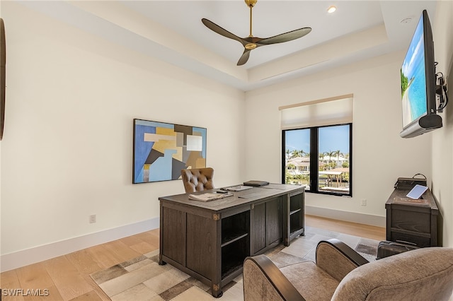 home office featuring ceiling fan, a tray ceiling, and light wood-type flooring