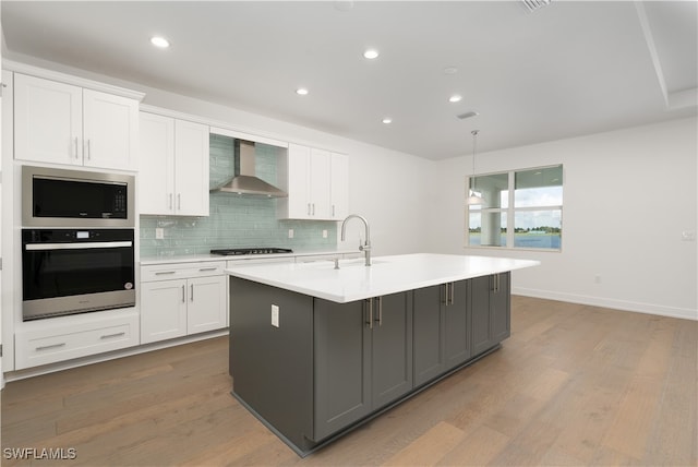 kitchen featuring wall chimney range hood, a center island with sink, hardwood / wood-style floors, white cabinets, and appliances with stainless steel finishes