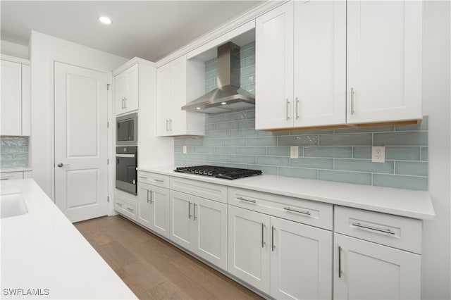 kitchen with wall chimney exhaust hood, stainless steel appliances, wood-type flooring, white cabinetry, and tasteful backsplash