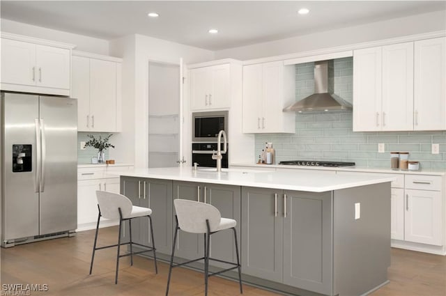 kitchen featuring stainless steel appliances, wall chimney exhaust hood, an island with sink, and white cabinets