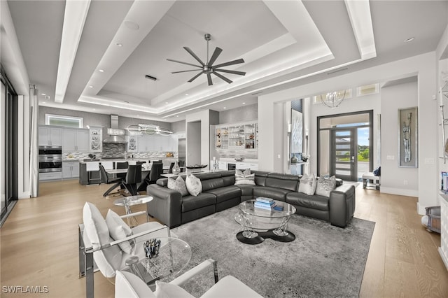 living room with ceiling fan with notable chandelier, a tray ceiling, and light wood-type flooring