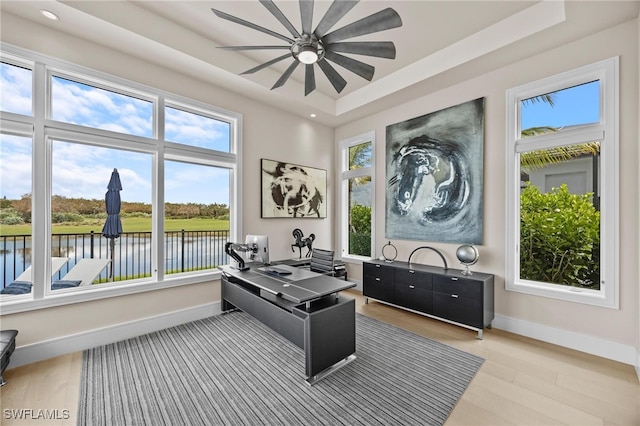 office space featuring light hardwood / wood-style flooring, a water view, a tray ceiling, and ceiling fan