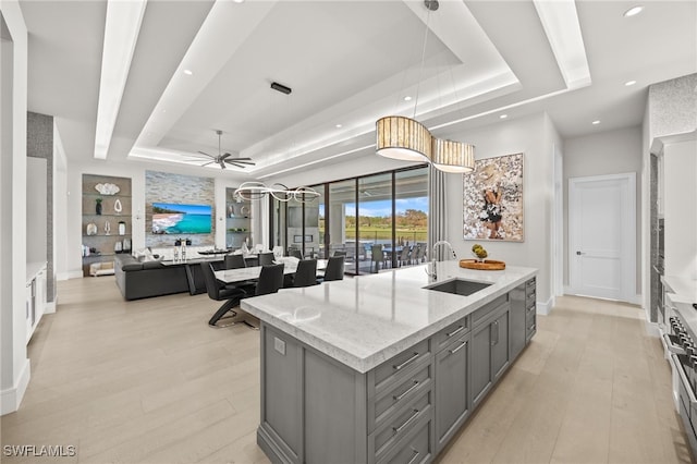 kitchen with an island with sink, a tray ceiling, pendant lighting, gray cabinetry, and sink