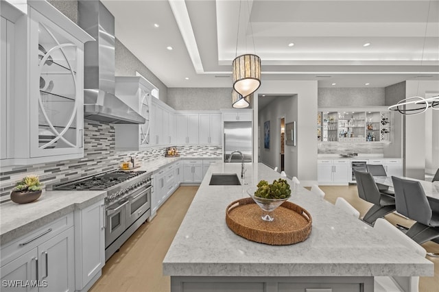 kitchen featuring a large island with sink, light wood-type flooring, pendant lighting, wall chimney exhaust hood, and premium appliances