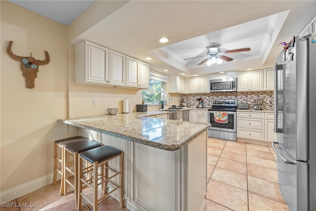 kitchen with appliances with stainless steel finishes, a tray ceiling, kitchen peninsula, light stone counters, and a breakfast bar area