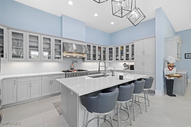 kitchen featuring wall chimney exhaust hood, sink, light tile patterned floors, decorative light fixtures, and an island with sink