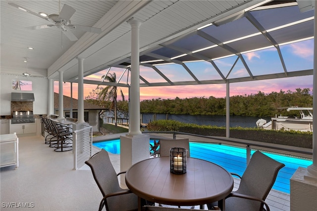 pool at dusk featuring grilling area, a water view, an outdoor bar, ceiling fan, and area for grilling