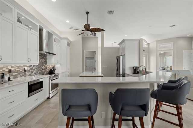 kitchen with ceiling fan, an island with sink, wall chimney range hood, and appliances with stainless steel finishes