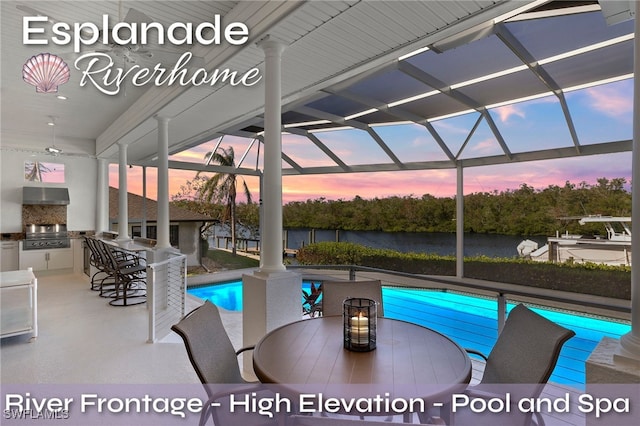 pool at dusk featuring glass enclosure, a water view, exterior bar, area for grilling, and exterior kitchen