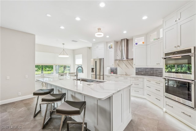 kitchen with white cabinetry, wall chimney range hood, pendant lighting, a center island with sink, and appliances with stainless steel finishes