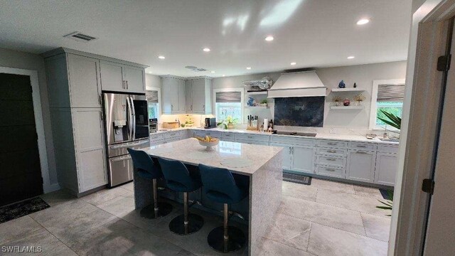 kitchen with a kitchen island, premium range hood, a kitchen bar, stainless steel refrigerator with ice dispenser, and light stone counters