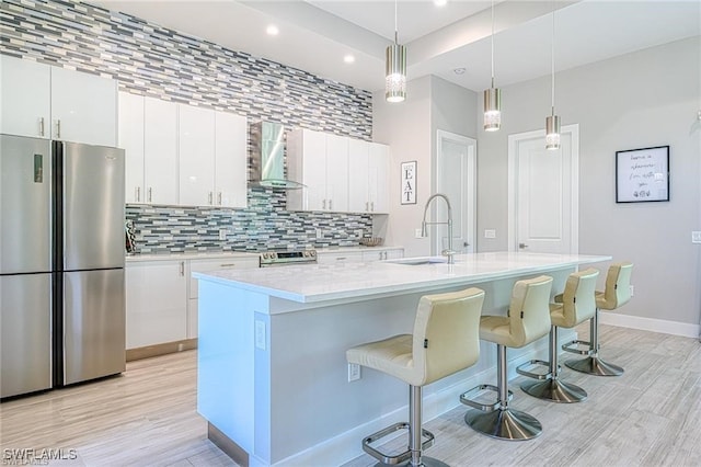 kitchen with wall chimney range hood, appliances with stainless steel finishes, sink, white cabinetry, and a kitchen island with sink