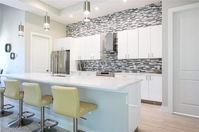 kitchen with wall chimney range hood, hanging light fixtures, a kitchen island with sink, sink, and stainless steel appliances