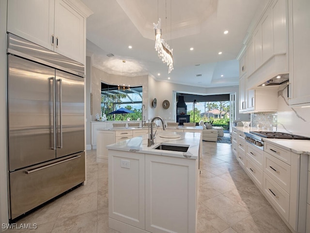 kitchen with a kitchen island with sink, a sink, a tray ceiling, appliances with stainless steel finishes, and decorative backsplash