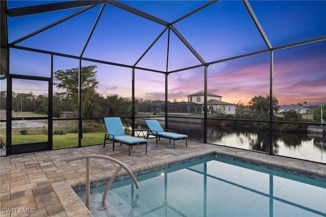 pool at dusk with glass enclosure, a patio area, and a water view