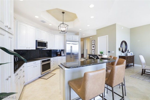 kitchen with stainless steel appliances, a tray ceiling, a center island with sink, white cabinetry, and hanging light fixtures