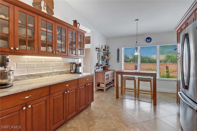 kitchen with decorative backsplash, hanging light fixtures, light tile patterned floors, light stone counters, and stainless steel refrigerator