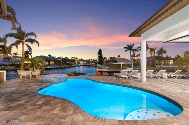 pool at dusk with an in ground hot tub, a water view, and a patio area