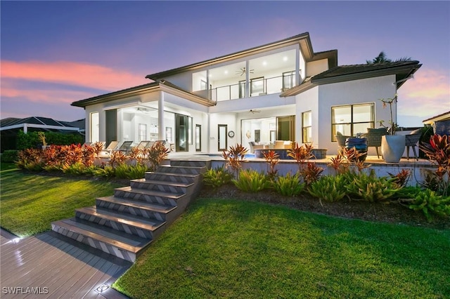 back house at dusk with a patio, a balcony, and a lawn