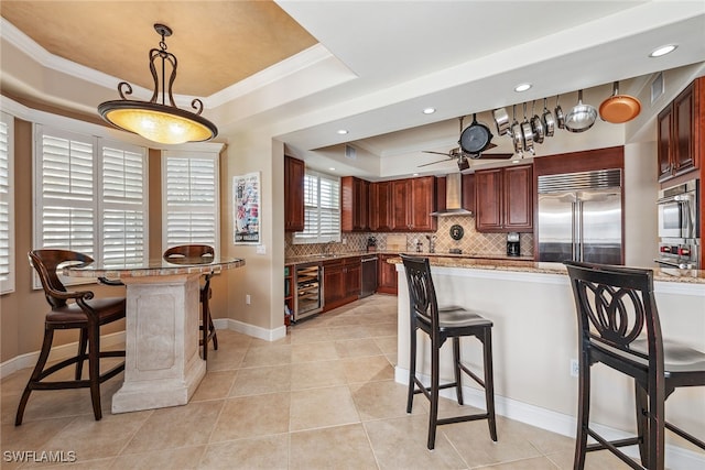 kitchen featuring stainless steel appliances, wine cooler, wall chimney range hood, pendant lighting, and a tray ceiling