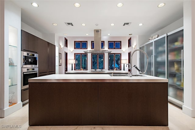 kitchen with dark brown cabinets, a center island with sink, double oven, and light tile patterned floors