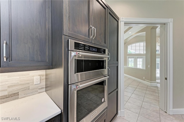 kitchen featuring stainless steel double oven, dark brown cabinets, crown molding, light tile patterned floors, and tasteful backsplash