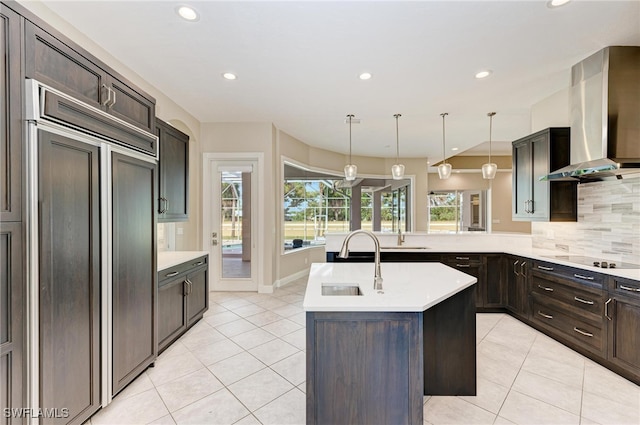 kitchen featuring paneled fridge, a center island with sink, hanging light fixtures, black electric cooktop, and wall chimney exhaust hood