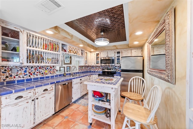 kitchen featuring decorative backsplash, tile counters, white cabinets, a raised ceiling, and appliances with stainless steel finishes