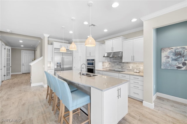 kitchen with light wood-type flooring, stainless steel appliances, a kitchen island with sink, pendant lighting, and white cabinets