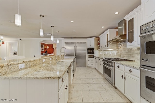 kitchen featuring built in appliances, white cabinetry, hanging light fixtures, and wall chimney range hood