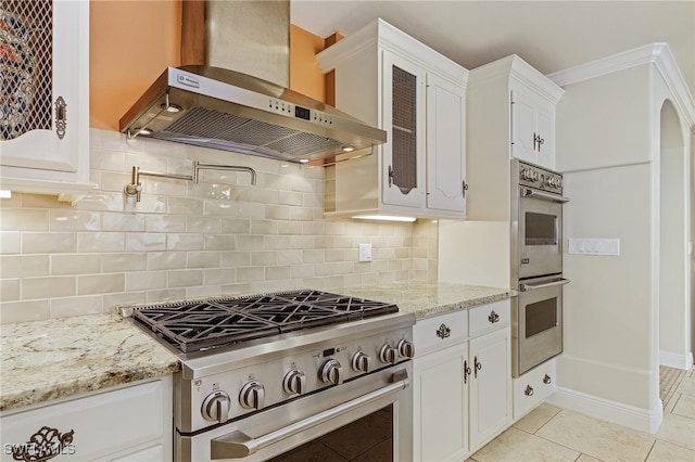 kitchen with backsplash, wall chimney range hood, appliances with stainless steel finishes, light tile patterned flooring, and white cabinetry