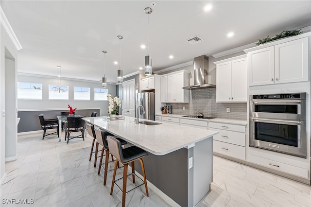 kitchen featuring stainless steel appliances, hanging light fixtures, a kitchen island with sink, crown molding, and wall chimney range hood