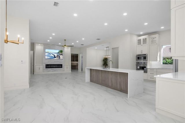 kitchen with a kitchen island with sink, hanging light fixtures, light stone countertops, a fireplace, and white cabinetry